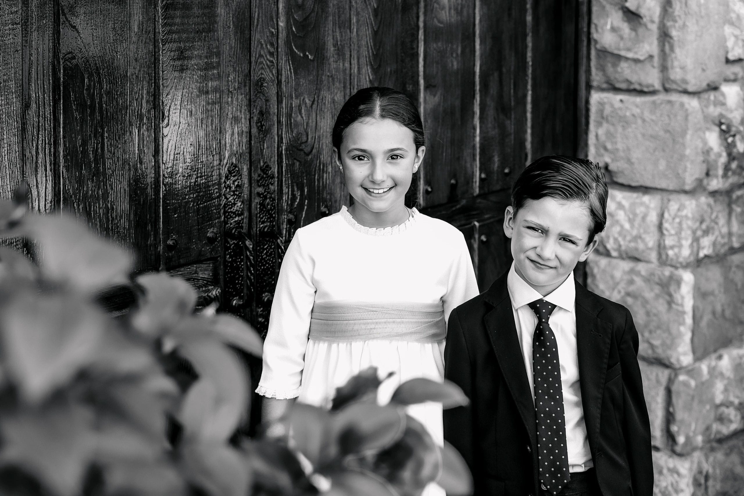 Niña y niño de comunión ante madera y piedra, en blanco y negro