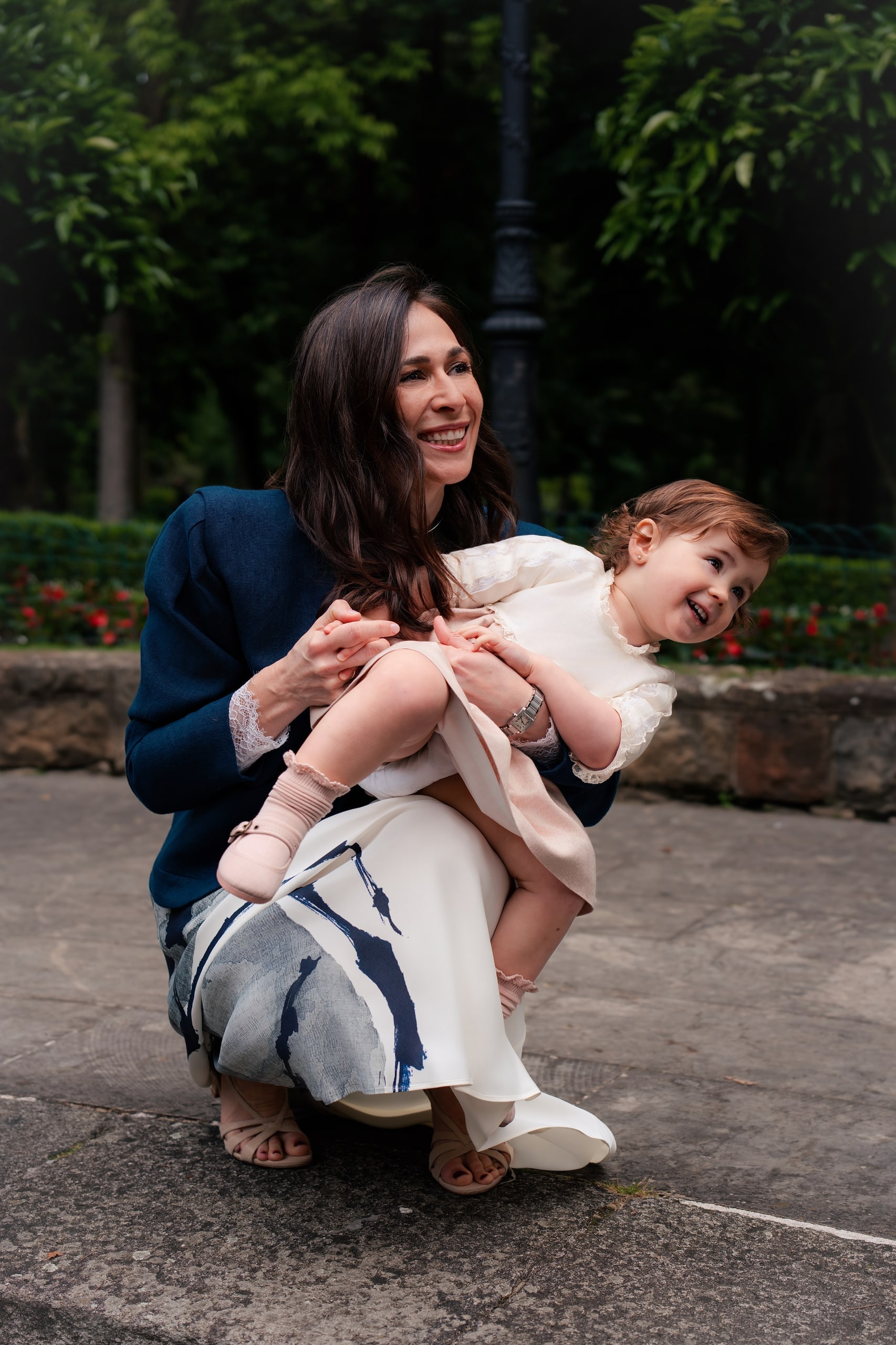 Una madre, en cuclillas en el parque, sostiene a su hija pequeña sobre el regazo; las dos se ríen, mirando cada una hacia un lado