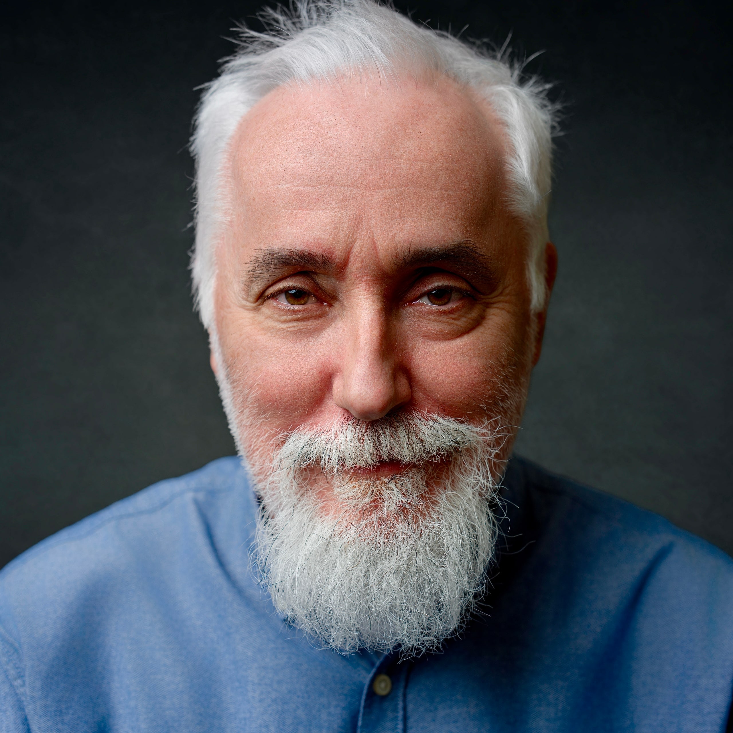 Retrato de estudio de un hombre de barba blanca y camisa azul, mirando a cámara